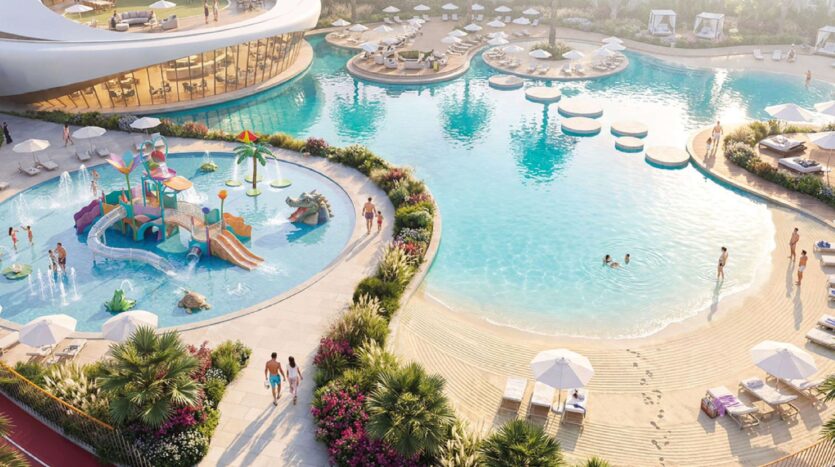 Luftaufnahme des Pools des Greenz by Danube-Komplexes mit einem Sandstrand am Eingang, Liegestühlen, Sonnenschirmen und einem bunten Kinderspielplatz. Mehrere Personen baden und entspannen sich, umgeben von üppigen Pflanzen und Gärten.