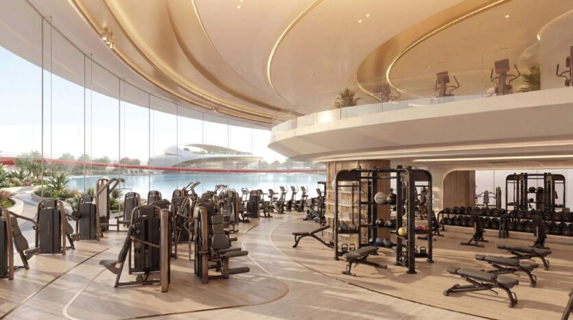 Geräumiger und moderner Fitnessraum im Greenz by Danube mit großen Fenstern, viel Tageslicht, verschiedenen Trainingsgeräten, Hanteln und einer offenen, geschwungenen Raumaufteilung mit einer oberen Ebene. Von außen ist ein futuristisches Gebäude zu sehen.