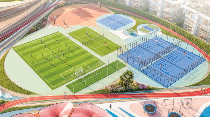 Luftaufnahme von „Greenz by Danube“, die einen farbenfrohen Sportkomplex mit einem Fußballplatz, Tennisplätzen, blauen Paddle-Plätzen, einer Laufbahn und Menschen zeigt, die aktiv Sport treiben oder trainieren. Moderne Gebäude umgeben die Sportplätze.
