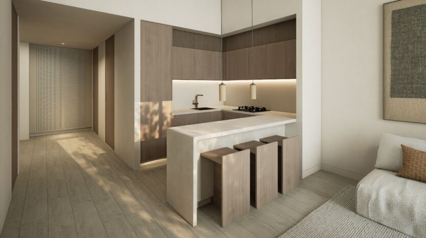Modern minimalist kitchen with wooden cabinets, a light-colored countertop, and three wooden bar stools. The well-lit space of unit JAD288 features neutral tones and opens onto a hallway and living area with a sofa and wall art.