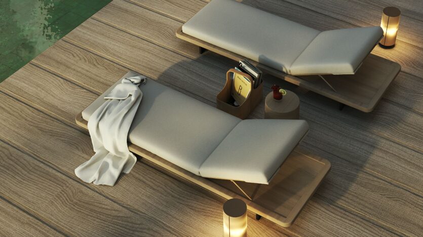 A modern JAD288 pool lounger, covered with a white towel, sits on a wooden deck next to a small table with drinks and books. Soft lighting and another chair are visible nearby.