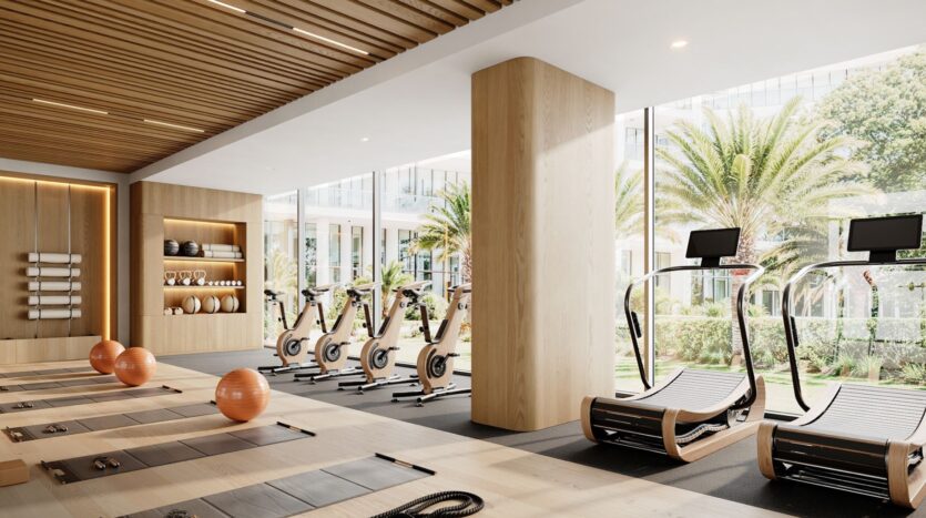 Salle de sport moderne au Tara Park avec de grandes fenêtres, des accents en bois, des tapis d'exercice, des balles de stabilité orange, des vélos stationnaires et des tapis roulants incurvés. La verdure luxuriante et les palmiers à l'extérieur créent une atmosphère sereine.