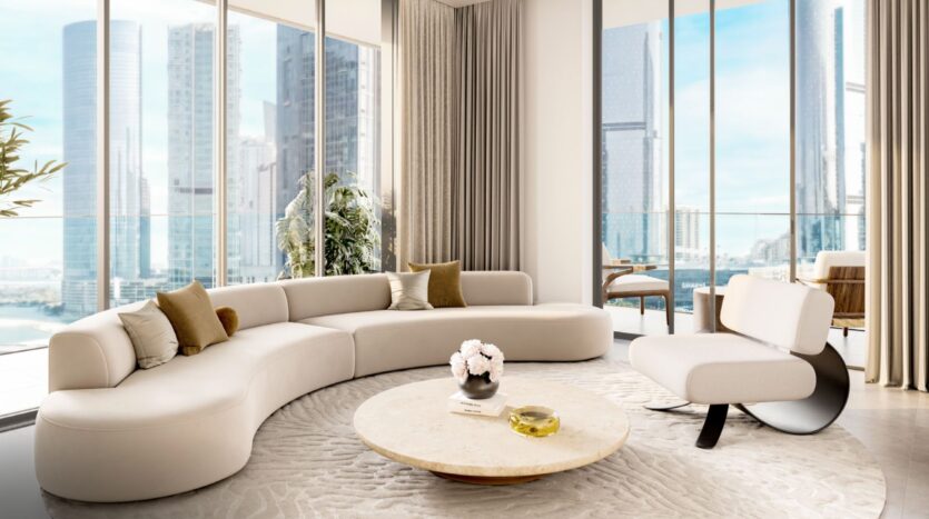 Un salon moderne à Tara Park comprend un canapé blanc incurvé, une table basse ronde, un fauteuil d'appoint et des fenêtres allant du sol au plafond avec vue sur le paysage urbain. Des tons neutres et une plante en pot ajoutent une touche de confort et d'élégance.