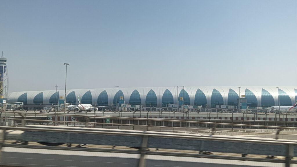 Am Flughafen Dubai erhebt sich unter klarem Himmel ein großes, modernes Flughafenterminal mit einem geschwungenen blau-weißen Design, vor dem mehrere Flugzeuge im Freien stehen und links der Flugsicherungsturm zu sehen ist.