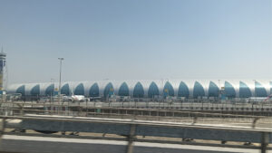 Am Flughafen Dubai erhebt sich unter klarem Himmel ein großes, modernes Flughafenterminal mit einem geschwungenen blau-weißen Design, vor dem mehrere Flugzeuge im Freien stehen und links der Flugsicherungsturm zu sehen ist.