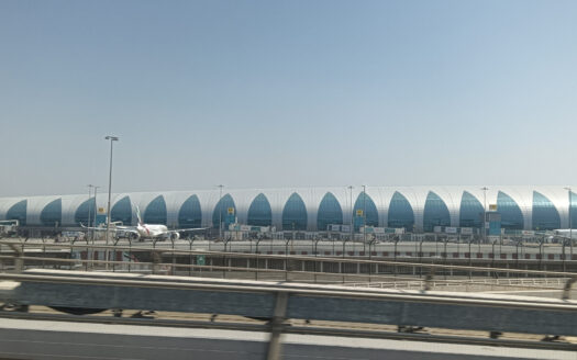 Un grand terminal aéroportuaire moderne avec un design arqué bleu et blanc se dresse sous un ciel clair à l'Aéroport de Dubaï, avec plusieurs vols garés à l'extérieur et une tour de contrôle du trafic aérien visible sur la gauche.