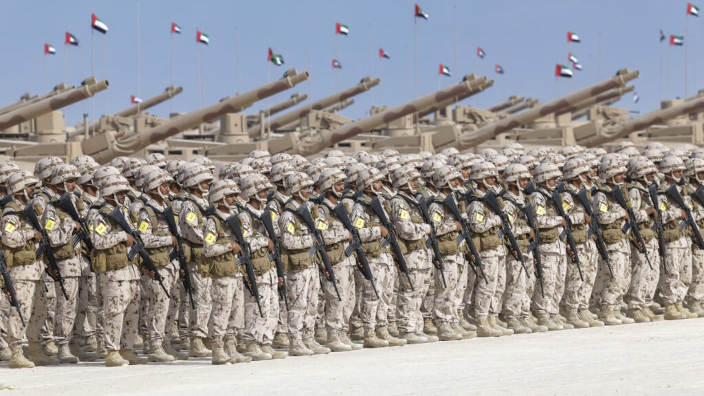 A large formation of soldiers in camouflage uniforms stands in ranks on a parade ground, illustrating the military might of the United Arab Emirates, with tanks and artillery behind them and national flags flying under the blue Gulf sky.