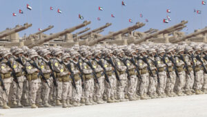 Eine große Formation von Soldaten in Tarnuniformen steht in Reih und Glied auf einem Exerzierplatz und demonstriert die militärische Stärke der Vereinigten Arabischen Emirate, hinter ihnen Panzer und Artillerie und unter dem blauen Himmel des Golfs wehende Nationalflaggen.