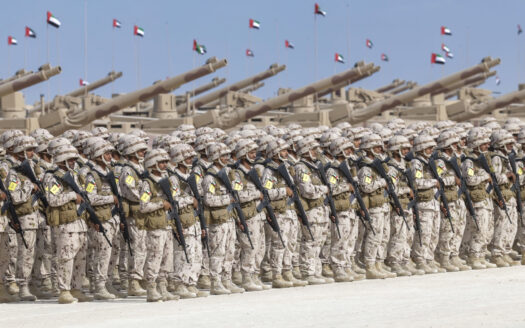 Eine große Formation von Soldaten in Tarnuniformen steht in Reih und Glied auf einem Exerzierplatz und demonstriert die militärische Stärke der Vereinigten Arabischen Emirate, hinter ihnen Panzer und Artillerie und unter dem blauen Himmel des Golfs wehende Nationalflaggen.