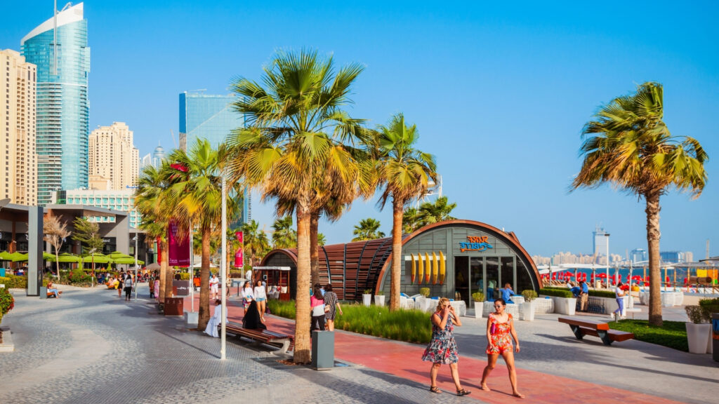In una giornata di sole, la gente passeggia lungo una passeggiata fiancheggiata da palme, con edifici moderni e negozi sullo sfondo; alcuni si siedono sulle panchine, mentre altri bighellonano o chiacchierano, discutendo di come gli abitanti di Dubai stiano vivendo la crisi legata alla guerra in Iran.