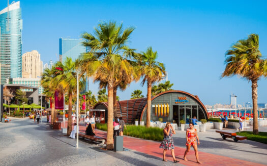 An einem sonnigen Tag spazieren Menschen eine von Palmen gesäumte Promenade entlang, im Hintergrund moderne Gebäude und Geschäfte; einige sitzen auf Bänken, während andere umherflanieren oder sich unterhalten und darüber diskutieren, wie die Einwohner Dubais die Krise im Zusammenhang mit dem Krieg im Iran erleben.
