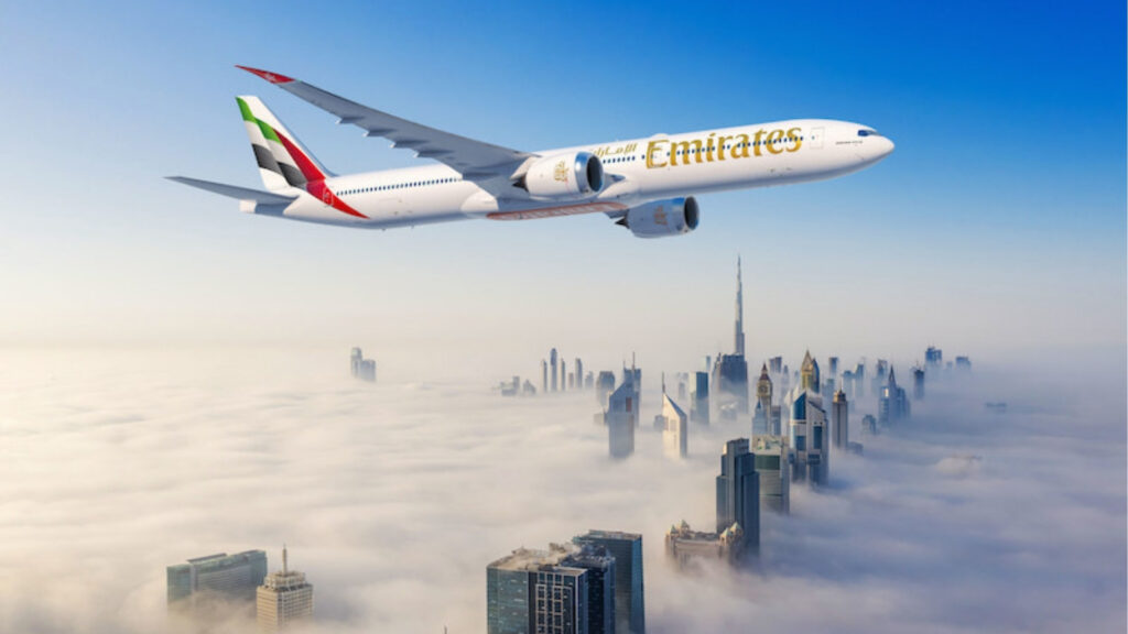 Самолет авиакомпании Emirates пролетает над городским пейзажем, окутанным туманом, из которого вырисовываются такие знаковые небоскребы, как Бурдж-Халифа, символизируя ту уверенность, которую Дубай дарит тем, кто хочет посетить этот город под ясным голубым небом.