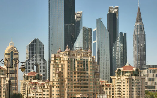 Blick auf die Skyline mit beigefarbenen, verzierten Wohngebäuden im Vordergrund und modernen Glaswolkenkratzern im Hintergrund unter strahlend blauem Himmel, die die Widerstandsfähigkeit Dubais inmitten der Immobilienkrise und globaler Spannungen wie dem Krieg im Iran widerspiegeln.