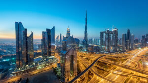 Luftaufnahme der Skyline von Dubai in der Abenddämmerung mit modernen Wolkenkratzern wie dem Burj Khalifa. Ein inspirierender Anblick für Investoren, die auch in Krisenzeiten nach Stabilität suchen, mit beleuchteten Autobahnen und einem klaren blauen Himmel, der in die Nacht übergeht.
