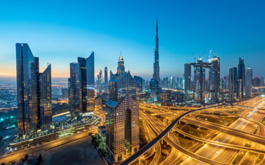 Luftaufnahme der Skyline von Dubai in der Abenddämmerung mit modernen Wolkenkratzern wie dem Burj Khalifa. Ein inspirierender Anblick für Investoren, die auch in Krisenzeiten nach Stabilität suchen, mit beleuchteten Autobahnen und einem klaren blauen Himmel, der in die Nacht übergeht.