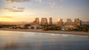 Mitten im Herzen von Dubais Immobilienmarkt erstrahlt ein moderner Strandkomplex mit seinen Glastürmen und üppiger Vegetation im goldenen Licht des Sonnenuntergangs, das sich im ruhigen Wasser spiegelt. Entlang des friedlichen Ufers säumen Sonnenliegen den Sandstrand.