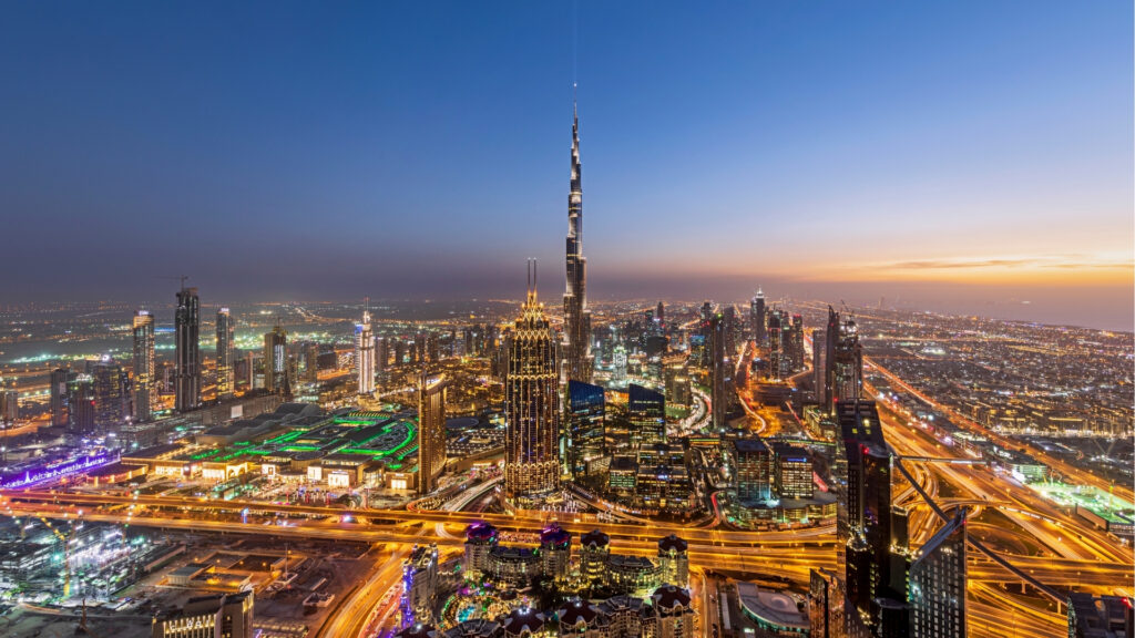An aerial view of the city of Dubai at dusk, featuring a brightly lit skyline and the Burj Khalifa towering above the skyscrapers, along with busy highways, illustrating Dubai’s vibrant energy and the business opportunities expected to grow significantly by 2026.