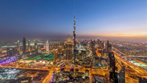 Luftaufnahme der Stadt Dubai in der Abenddämmerung, mit der hell erleuchteten Skyline und dem Burj Khalifa, der die Wolkenkratzer überragt, sowie stark befahrenen Autobahnen, die die pulsierende Energie Dubais und die Geschäftsmöglichkeiten verdeutlichen, die sich bis 2026 voraussichtlich vervielfachen werden.