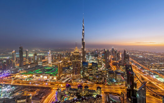 An aerial view of the city of Dubai at dusk, featuring a brightly lit skyline and the Burj Khalifa towering above the skyscrapers, along with busy highways, illustrating Dubai’s vibrant energy and the business opportunities expected to grow significantly by 2026.