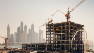 Ein im Bau befindliches Hochhaus mit Kränen ragt aus der nebligen Skyline von Dubai heraus. Sand, Baumaterialien und Geräte füllen den Vordergrund und verdeutlichen die sich bietenden Immobilienchancen in dieser dynamischen Stadtlandschaft.
