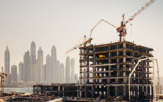 Ein im Bau befindliches Hochhaus mit Kränen ragt aus der nebligen Skyline von Dubai heraus. Sand, Baumaterialien und Geräte füllen den Vordergrund und verdeutlichen die sich bietenden Immobilienchancen in dieser dynamischen Stadtlandschaft.