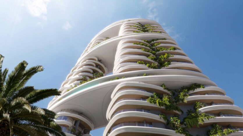 Amali Residences es un moderno rascacielos con balcones curvos, cada uno bordeado de exuberante vegetación, que se recortan contra un cielo azul. En primer plano, a la izquierda, se ve una palmera.