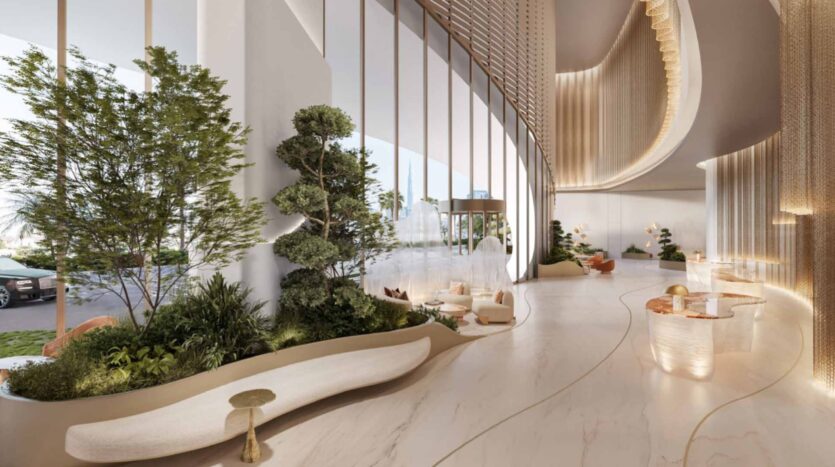 El vestíbulo del hotel Amali Residences es moderno y espacioso, con ventanas altas, detalles dorados curvados, plantas en el interior, suelos de mármol claro, cómodos asientos y una suave iluminación natural. A través de las ventanas se divisan un coche y unos árboles al aire libre.