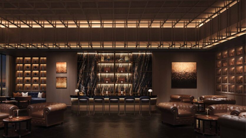 Un sofisticado bar-salón en las residencias Amali, con sofás y sillones de cuero marrón, una barra de mármol con taburetes, una cálida iluminación ambiental, obras de arte abstractas en las paredes y una pared con estanterías en la que se exponen botellas y objetos decorativos.