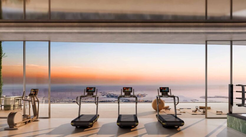 Tres cintas de correr se encuentran frente a grandes ventanales que van del suelo al techo y ofrecen unas vistas panorámicas del mar y de la ciudad al atardecer, en un moderno gimnasio de las residencias Amali, dotado de un equipamiento elegante y un ambiente sereno y lujoso.