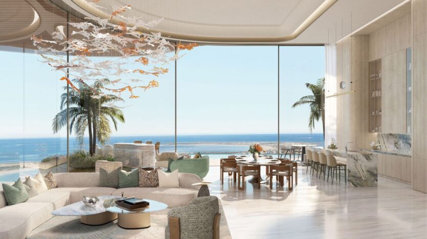 Un salón y un comedor lujosos y modernos en las residencias Amali, con ventanas de suelo a techo con vistas al océano, suelos de mármol, muebles en tonos neutros, una lámpara de araña abstracta y palmeras visibles desde el exterior.