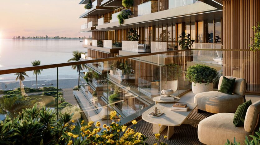 A luxurious balcony at the Cheval Residences, featuring modern furnishings, a glass railing, potted plants, and a coffee table, overlooking a tranquil beach and the ocean at sunset. Flowers and palm trees enhance the elegant and relaxing atmosphere.