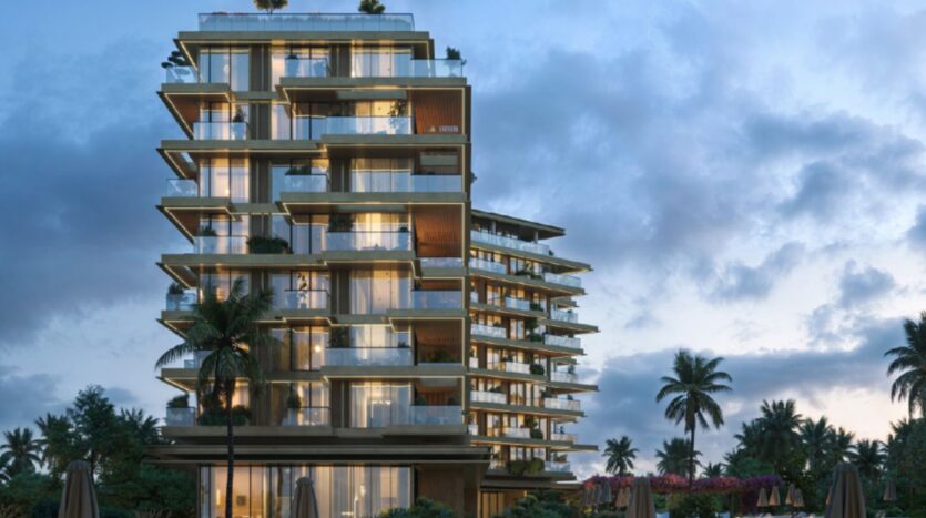 A modern multi-story building with glass balconies, illuminated at dusk, surrounded by palm trees and lush greenery under a cloudy sky—an iconic setting at Cheval Residences.
