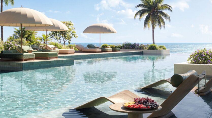 An infinity pool overlooking the ocean at Les Résidences Cheval, complete with lounge chairs, umbrellas, and a platter of fresh fruit. Lush greenery and palm trees surround the pool, creating a relaxing tropical atmosphere under a clear, bright sky.