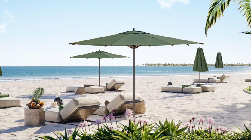 A sunny beach scene at the Cheval Residences, featuring lounge chairs and green umbrellas on the white sand, facing a calm blue ocean. Tropical plants and flowers fill the foreground, while a tray of fruit and drinks sits on a small table.