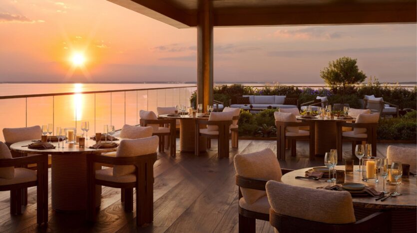 The elegant terrace of the Cheval Residences’ outdoor restaurant overlooks a calm body of water at sunset, featuring set tables, upholstered chairs, a glass railing, and soft, warm lighting that creates a serene and inviting atmosphere.