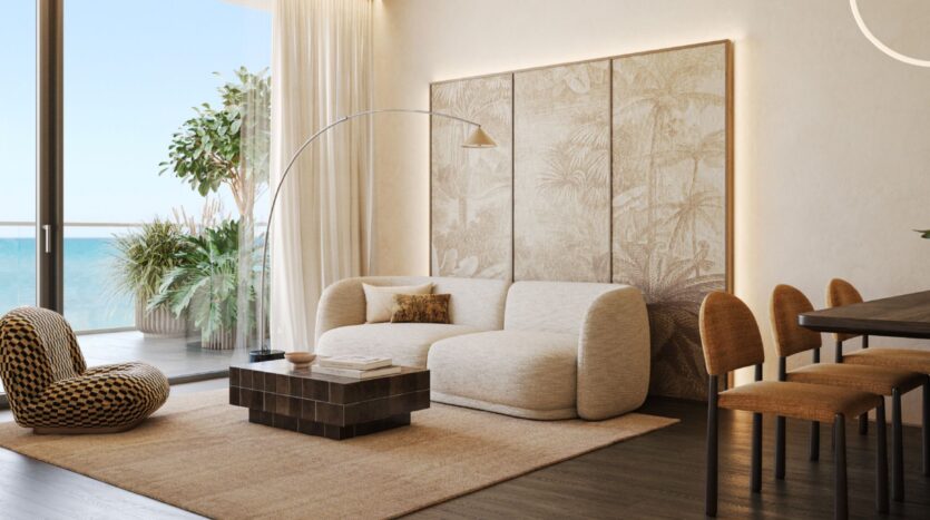 The modern living room at Cheval Residences features a cream-colored sofa, a patterned armchair, and a dark wood coffee table. Large windows offer ocean views, while neutral tones and wall art create a calm and airy atmosphere.