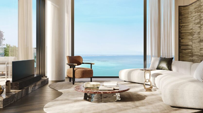 A modern living room at Les Résidences Cheval, featuring large windows that offer a panoramic view of the ocean. The space features a curved white sofa, a brown armchair, a marble coffee table, and a neutral color scheme, with natural light flooding in.
