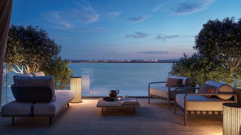 The outdoor patio at Cheval Residences features upholstered seating, a coffee table, and potted trees overlooking a tranquil pond at dusk, with soft lighting that creates a relaxing atmosphere.