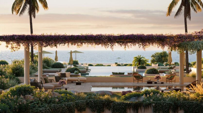 The luxurious outdoor lounge at the Cheval Residences, featuring sofas, chairs, and tables under a pergola covered in purple flowers, offers views of the pool, palm trees, gardens, and the ocean at sunset.