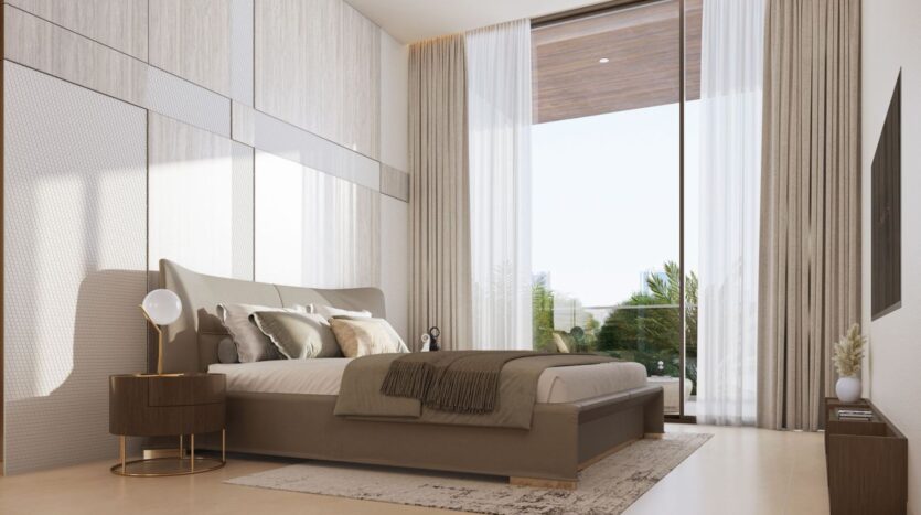 A modern bedroom in Derby Heights featuring a king-size bed, neutral bedding, and a round nightstand with a lamp. A floor-to-ceiling window with sheer curtains lets in natural light, offering a view of the greenery outside. The wall-mounted TV faces the bed.