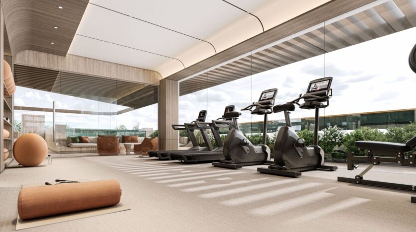 The modern fitness center at Derby Heights, featuring large windows, is equipped with treadmills, stationary bikes, exercise balls, yoga mats, and a seating area. The space is flooded with natural light, and greenery can be seen outside.