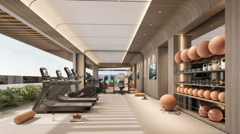 The modern gym at Derby Heights is equipped with treadmills, stationary bikes, and shelves stocked with kettlebells, yoga balls, and mats. Large windows flood the sleek, minimalist space with natural light and offer a glimpse of the lush greenery growing outside.