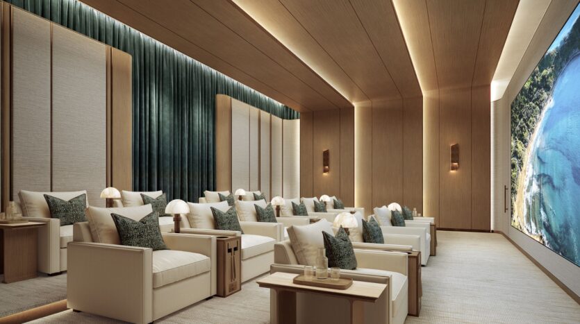 The luxurious movie theater at Four Seasons Private Residences features white armchairs, side tables with lamps, green curtains, wood-paneled walls and ceilings, and a large screen displaying a picturesque view of the coast.