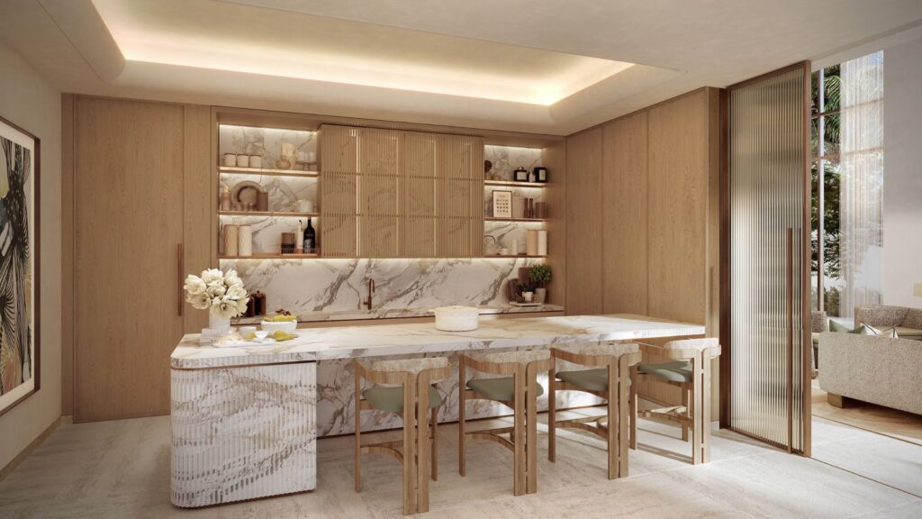 The modern kitchen at Four Seasons Private Residences features light wood cabinets, open shelving, a marble island with four wooden chairs, and built-in lighting. Decorative accents and fresh flowers create a clean and inviting atmosphere.