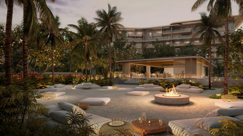 An outdoor lounge with low seating and a circular fire pit, surrounded by palm trees and lush greenery, set against the modern backdrop of the Four Seasons private residences and an open-air pavilion at sunset.