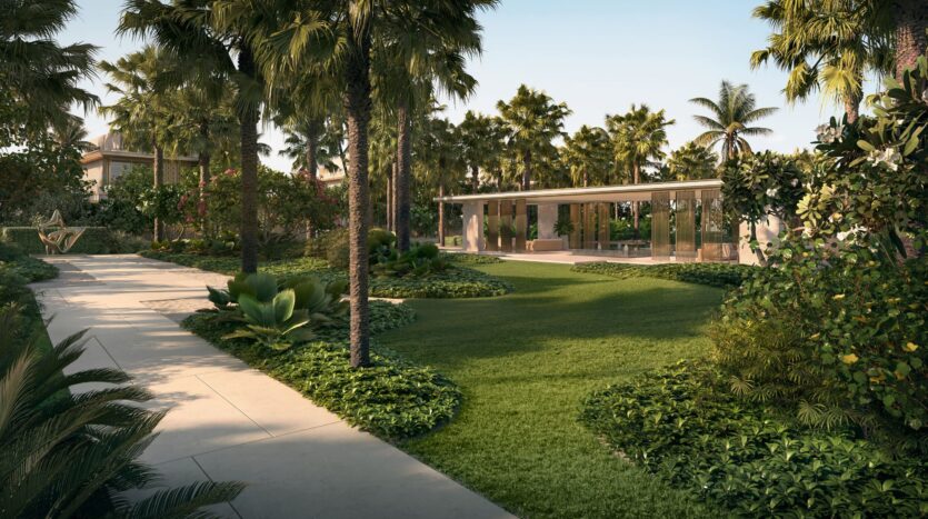 A lush tropical garden with palm trees, manicured lawns, and shrubs surrounds the Four Seasons private residences. Sunlight filters through the foliage along a winding paved path, leading to a modern glass-walled building in the background.