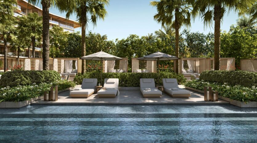 A luxurious poolside scene at Four Seasons Private Residences, featuring plush lounge chairs, umbrellas, and cabanas surrounded by lush greenery and palm trees, on a sunny day with a clear blue sky.