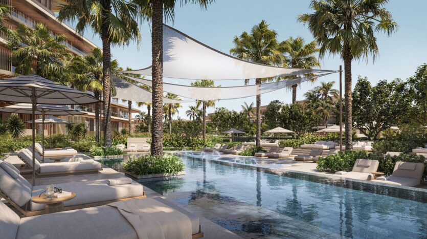 The luxurious outdoor pool at Four Seasons Private Residences, surrounded by lounge chairs, umbrellas, palm trees, and lush landscaping, with the resort’s modern buildings in the background, under a clear blue sky.