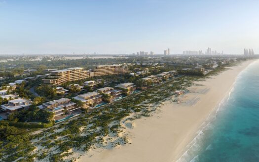 Luftaufnahme des Badeortes „Four Seasons Private Residences“, dessen moderne Gebäude von Grünflächen, einem Sandstrand und dem blauen Meer umgeben sind. Liegestühle und Sonnenschirme säumen das Ufer; im Hintergrund ist die Silhouette einer Stadt zu erkennen.