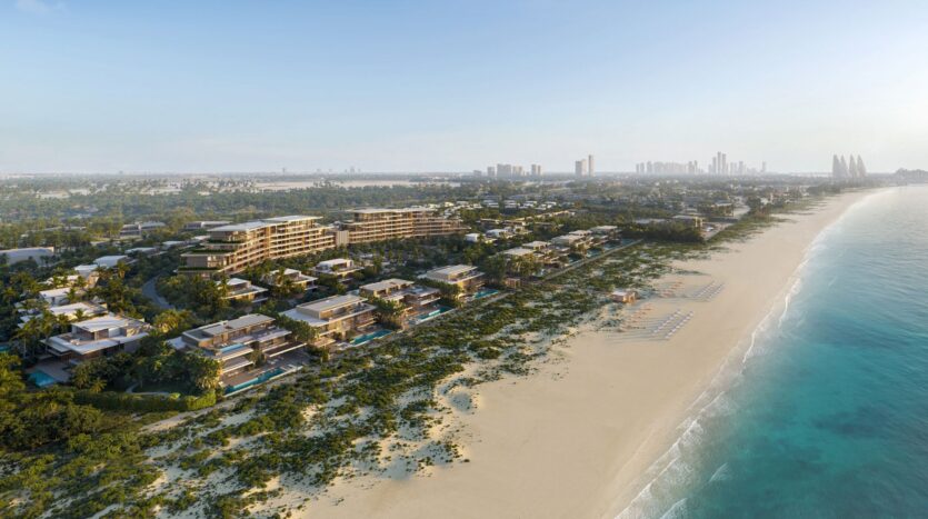 Aerial view of Four Seasons Private Residences, a luxury beach resort featuring modern buildings surrounded by lush greenery, facing a long sandy beach and turquoise waters, with the silhouette of a city visible in the background.